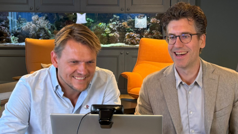 Twee mannen zitten aan een bureau met een laptop, omringd door een moderne inrichting.