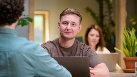 Man achter laptop in een moderne kantoorruimte met planten en laptops op de achtergrond.
