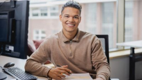 Man in beige trui werkt aan bureau bij raam met computer en documenten.