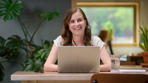 Vrouw achter laptop in lichte, groene ruimte met planten en groot raam.