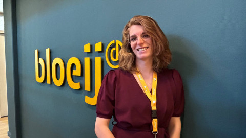 Vrouw in paarse jurk bij een muur met het logo "bloeij" in gele letters.