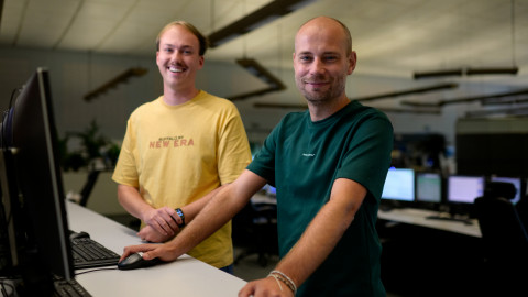 Twee personen staan bij een bureau in een moderne kantoorruimte met computerschermen op de achtergrond.