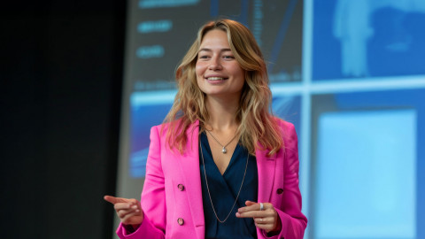 Vrouw in felroze pak spreekt met handgebaren, tegen een achtergrond van blauwe grafieken.