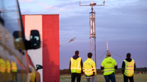 Mensen in gele hesjes kijken naar een vliegtuig bij een luchtvaarttoren in een open veld.