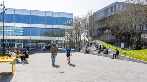 Een plein met moderne gebouwen, mensen zittend en wandelend, lentezon en groene grasrand.