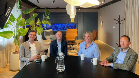 Vier mannen zitten aan een ronde tafel in een moderne ruimte met planten en een blauwe wanddecoratie.