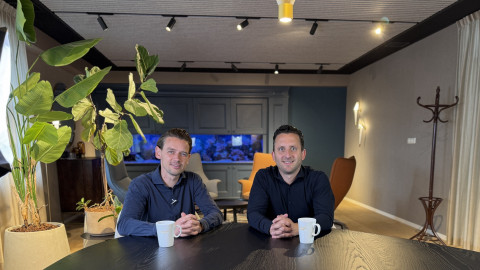 Twee mannen zitten aan een tafel met koffiekopjes in een moderne ruimte met groene planten.