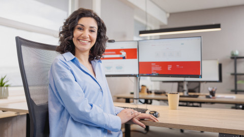 Vrouw glimlacht, zit aan een bureau met computerschermen en een kopje in een modern kantoor.