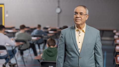 Man in een grijze jas, staand in een examenlokaal met studenten zittend aan tafels.