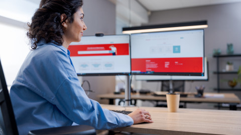 Vrouw aan bureau kijkt naar computerscherm met rood-witte interface in een modern kantoor.
