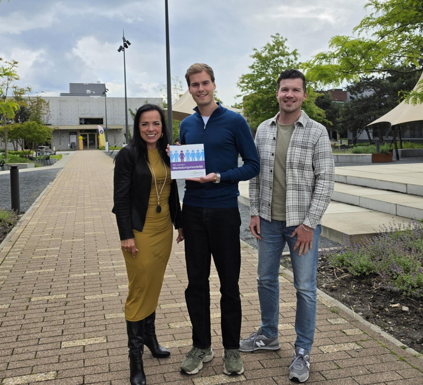 Drie mensen poseren buiten op een stenen pad; een man houdt een kleurrijke folder vast.