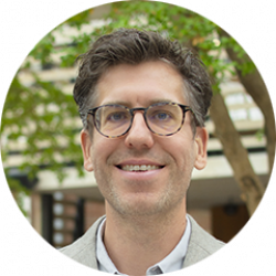 Man in grijze blazer met wit hemd, buiten met groene boom op achtergrond.