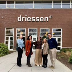 Vijf mensen staan glimlachend voor een gebouw met het logo "Driessen". 