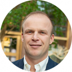 Man in een donkerblauw jasje en wit overhemd met groene planten op de achtergrond.