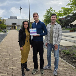 Drie mensen poseren buiten op een stenen pad; een man houdt een kleurrijke folder vast.