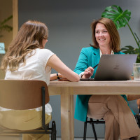 Twee vrouwen aan een tafel in gesprek, met een laptop en planten op de achtergrond.