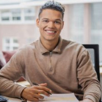 Man in beige trui werkt aan bureau bij raam met computer en documenten.