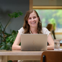 Vrouw achter laptop in lichte, groene ruimte met planten en groot raam.