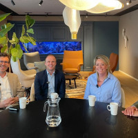 Vier mannen zitten aan een ronde tafel in een moderne ruimte met planten en een blauwe wanddecoratie.