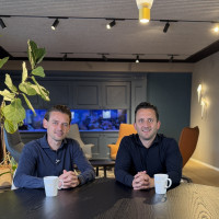 Twee mannen zitten aan een tafel met koffiekopjes in een moderne ruimte met groene planten.