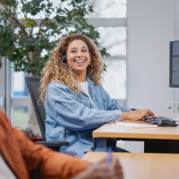 Vrouw met headset lacht terwijl ze aan bureau met computer werkt, man op de voorgrond vaag zichtbaar.