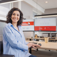 Vrouw glimlacht, zit aan een bureau met computerschermen en een kopje in een modern kantoor.