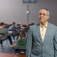 Man in een grijze jas, staand in een examenlokaal met studenten zittend aan tafels.